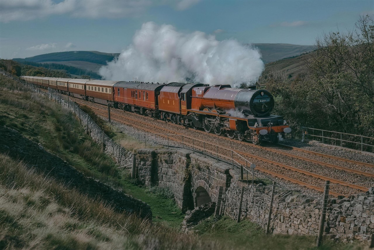 A red steam locomotive hauling a rake of carriages through open hill country